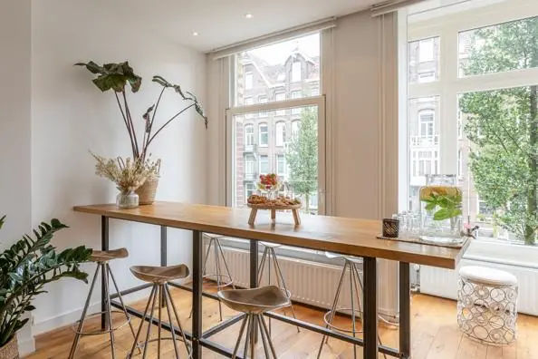 Hoge houten tafel met krukken in een licht interieur aan de Van Baerlestraat met uitzicht op Amsterdamse gevels.